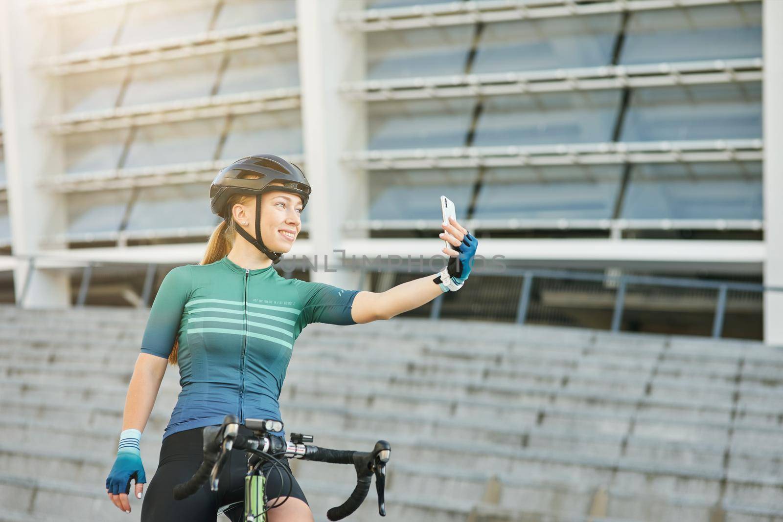 Adorable young woman, professional female cyclist smiling while holding smartphone and taking selfie, standing with her bike outdoors on a daytime by friendsstock