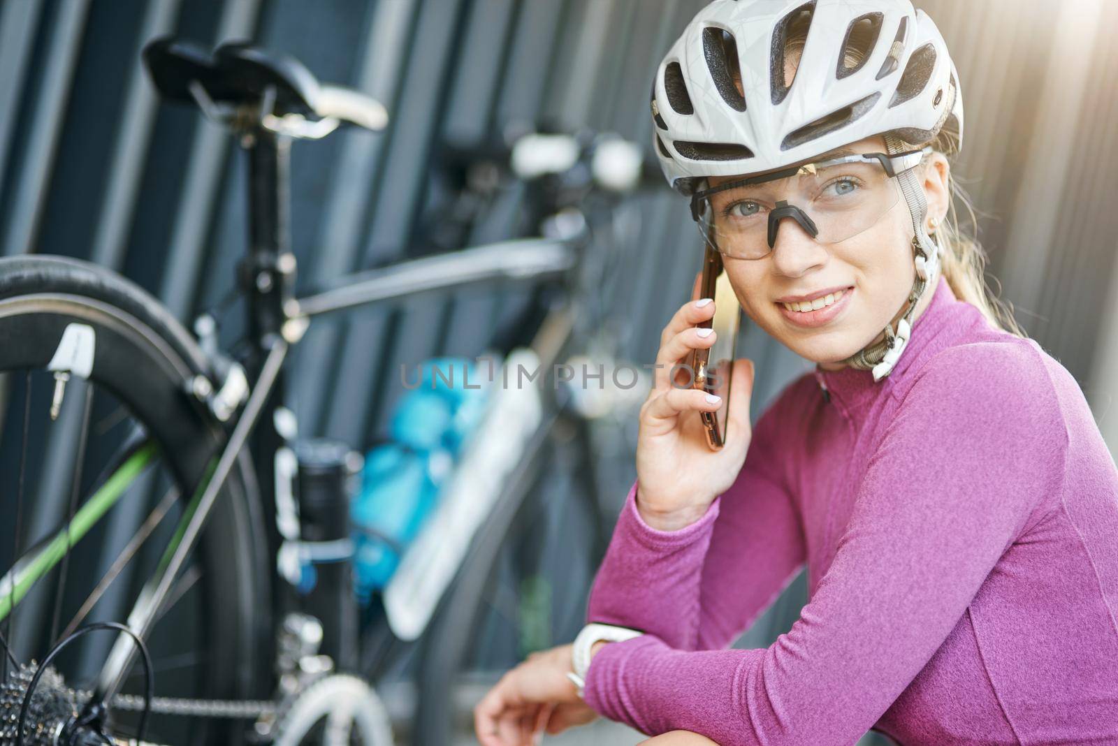 Portrait of attractive young sportive woman, female cyclist smiling, making a call using smartphone while checking her bike outdoors. Safety, sports, technology concept