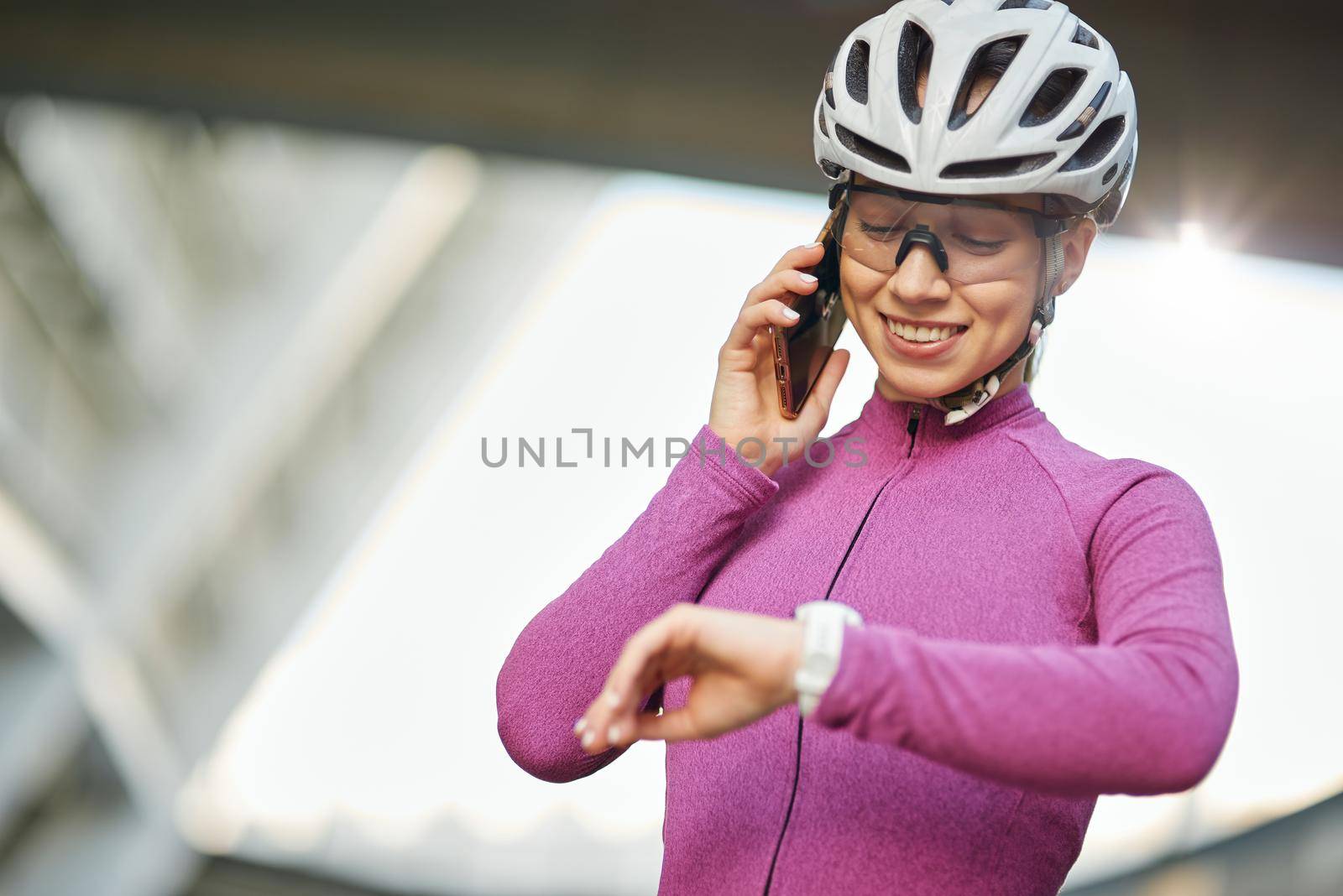 Cheerful young female cyclist wearing protective helmet and glasses looking at smartwatch on her wrist while talking on the phone, standing outdoors on a daytime. Safety, sports, technology concept