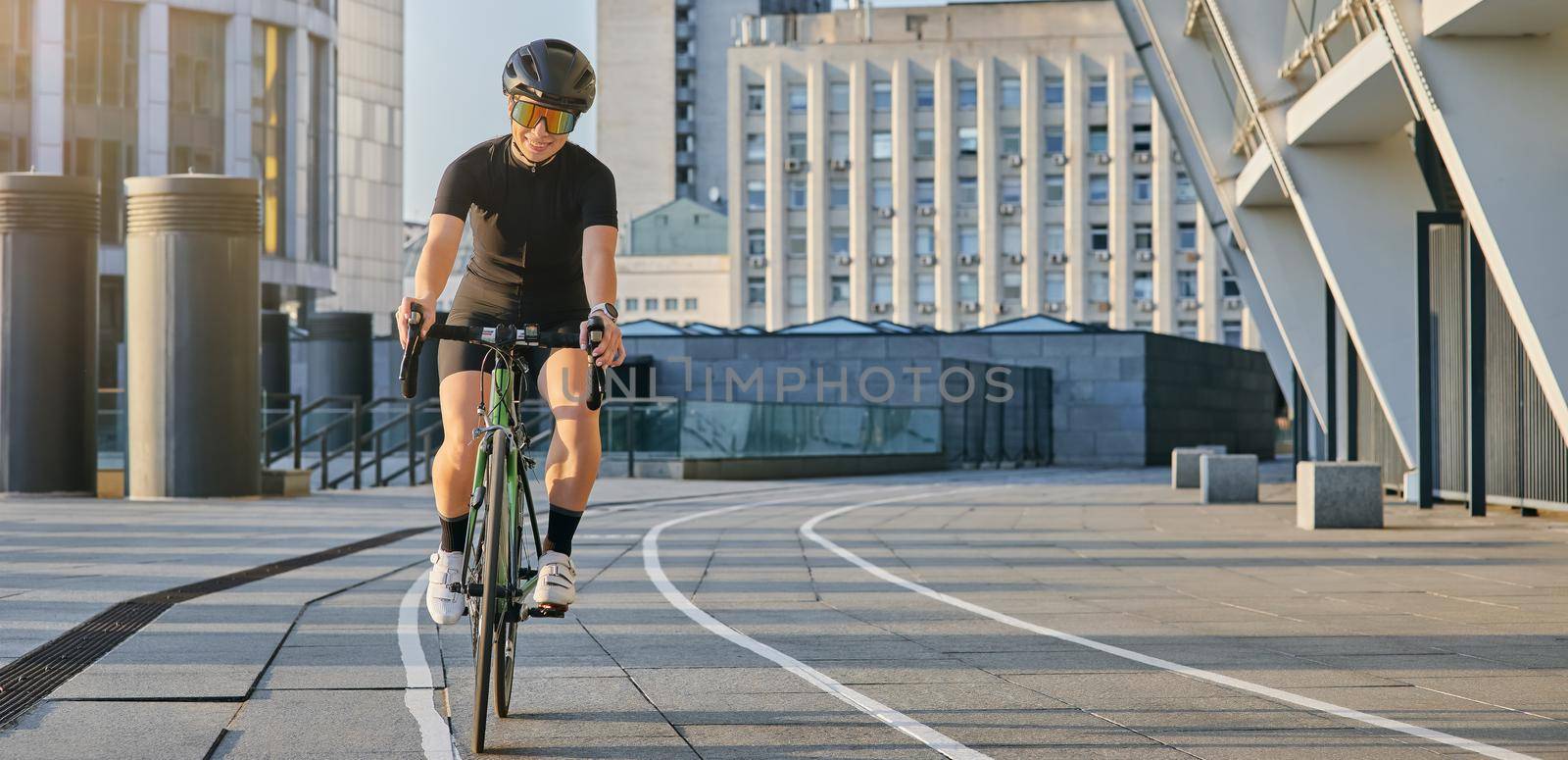 Delighted professional female cyclist in black cycling garment and protective gear smiling, looking satisfied with her training outdoors on a sunny day by friendsstock