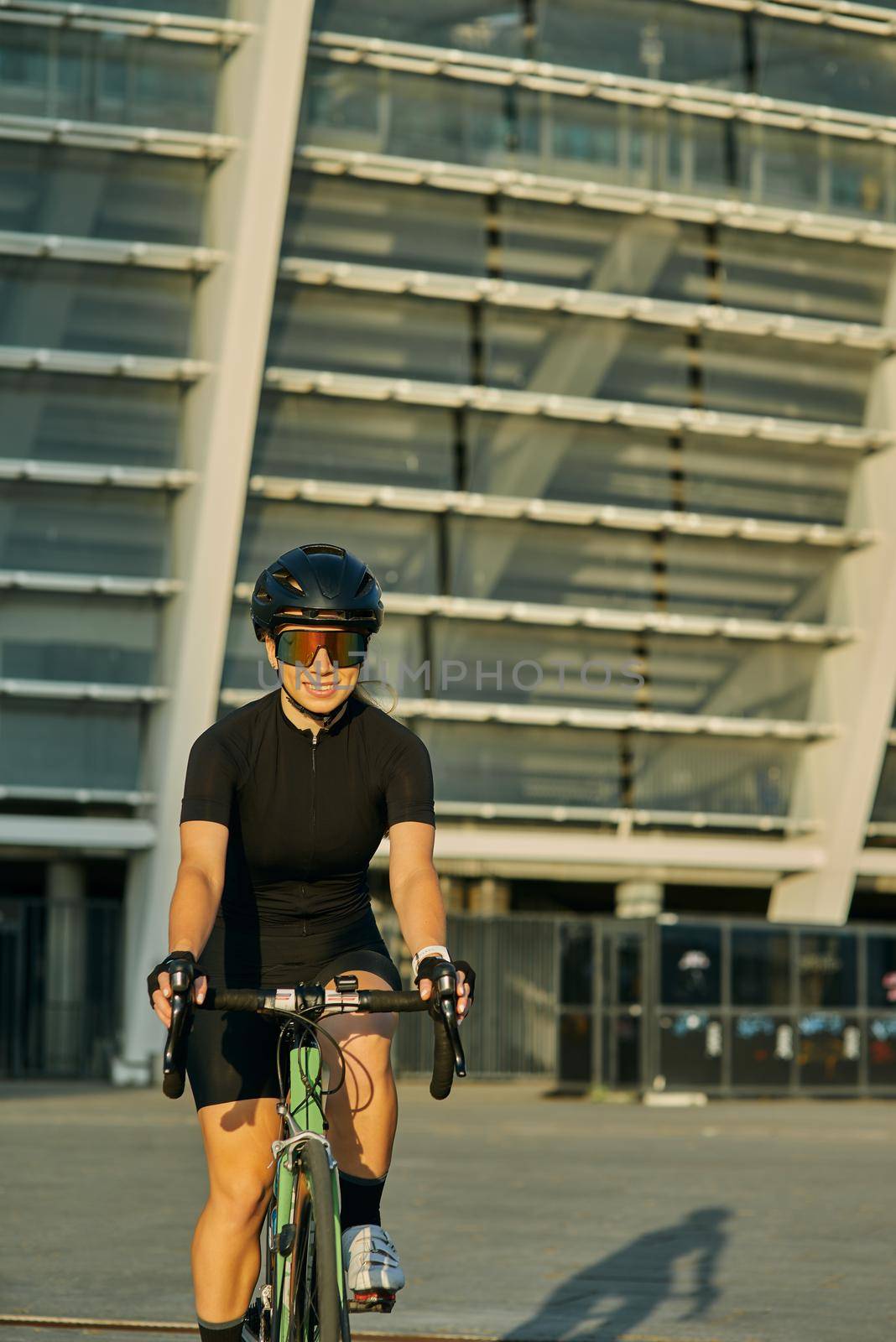 Excited professional female cyclist in black cycling garment and protective gear smiling while riding bicycle in city, training outdoors at sunset. Urban lifestyle, sports concept