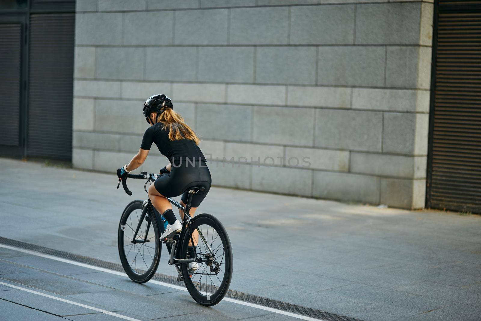 Rear view of professional female cyclist in black cycling garment and protective gear riding bicycle in city, passing buildings while training outdoors on a daytime. Urban lifestyle, sports concept