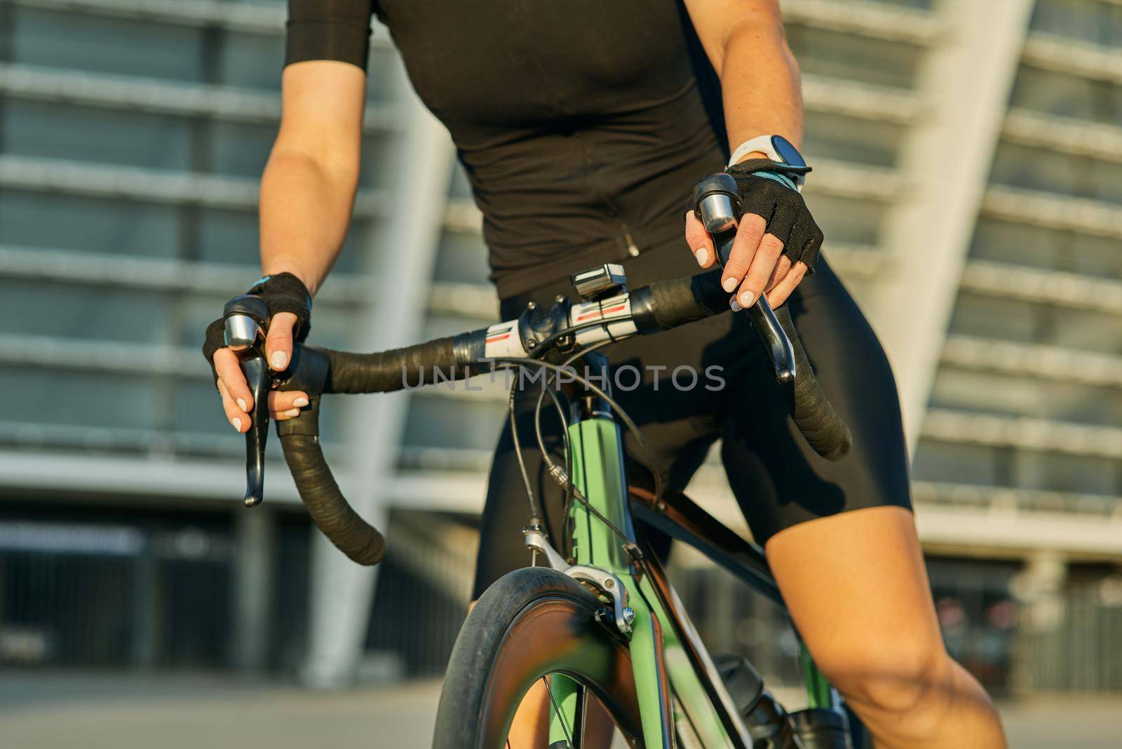Cropped shot of hands of female cyclist in black cycling garment and protective gear riding bike in city, training outdoors on a sunny day by friendsstock