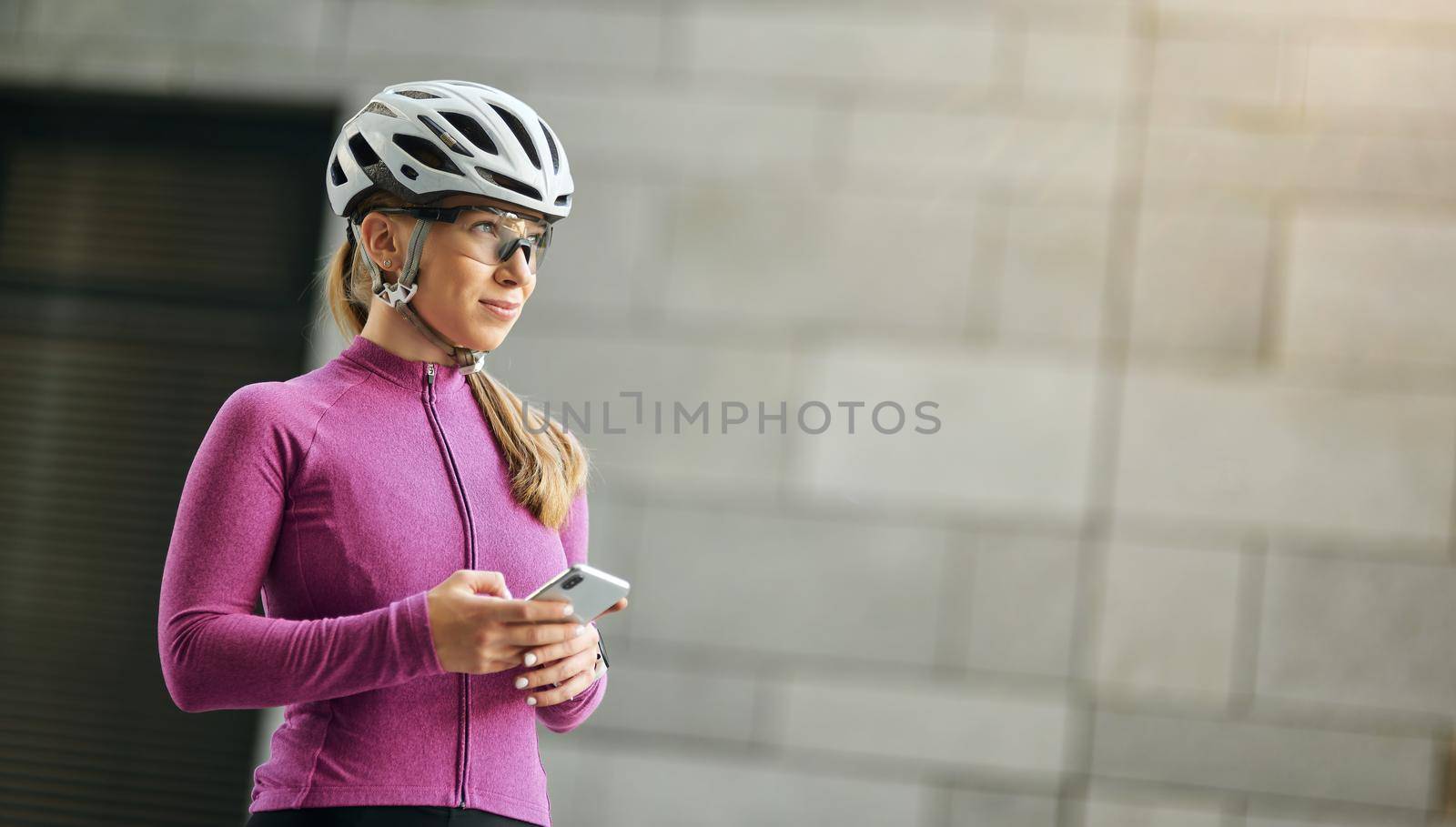 Professional female cyclist wearing pink suit and protective gear smiling away while holding smartphone, standing outdoors on a daytime. Sport, biking, people concept