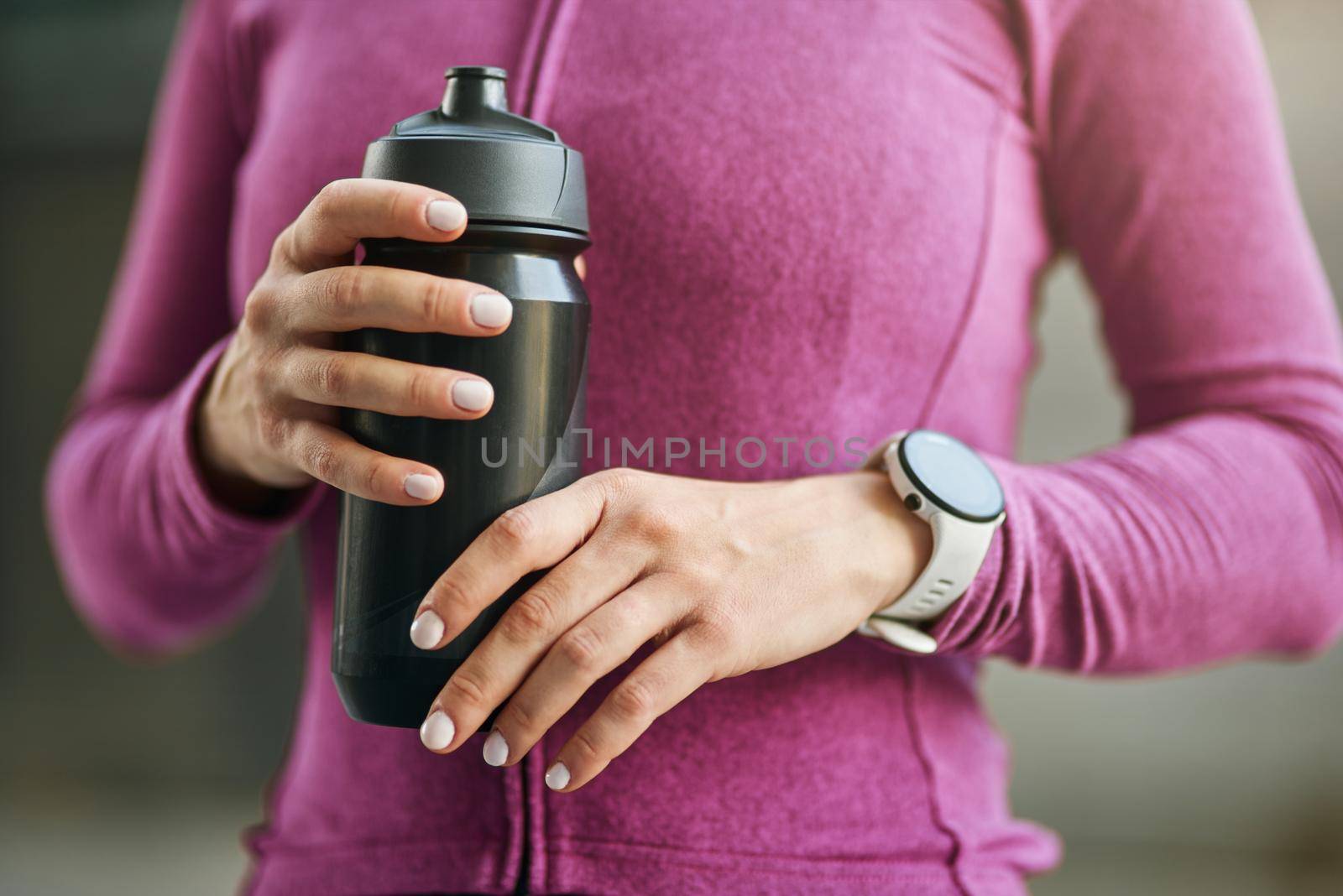 Close up of hands of sportive woman wearing pink suit holding bottle with drink while getting ready for training, standing outdoors on a daytime. Sports, active lifestyle concept
