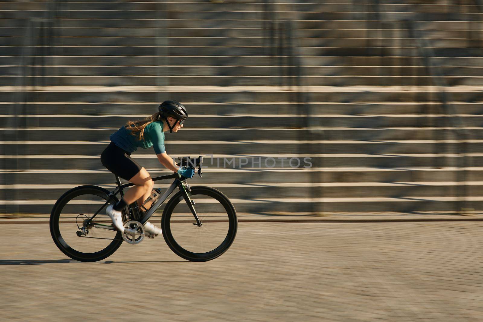 Side view of professional female cyclist in protective gear riding bicycle in city, rushing and passing buildings while training outdoors on a daytime. Urban lifestyle, sports concept
