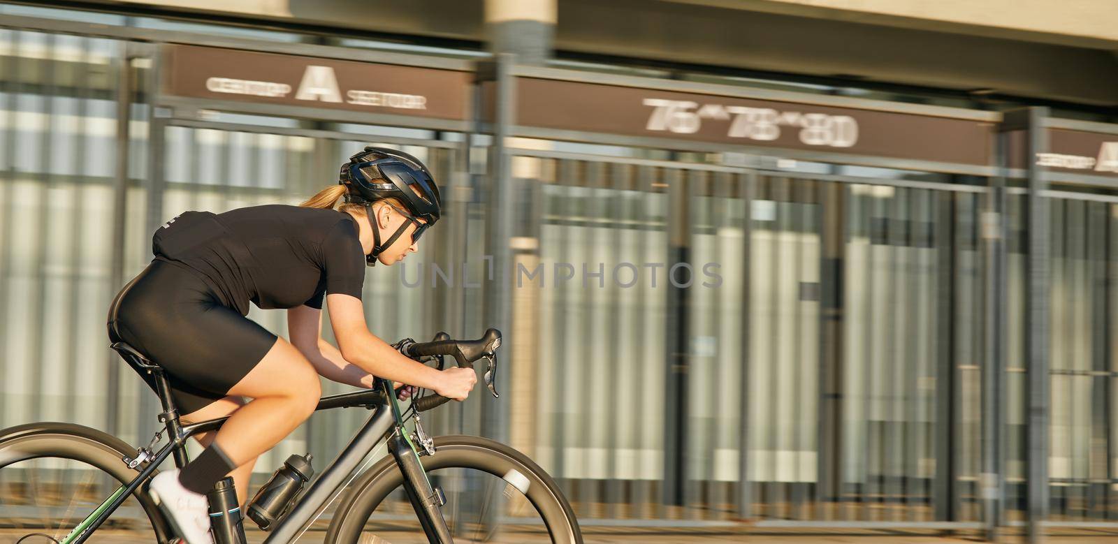Professional female cyclist in black cycling garment and protective gear riding bicycle in city center rushing and passing buildings by friendsstock