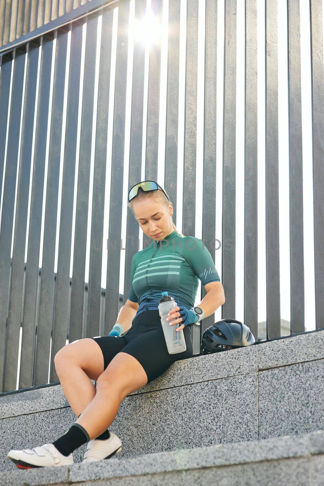 Young woman, professional cyclist in sporty shorts, with bottle of drink resting on the steps in the city background by friendsstock