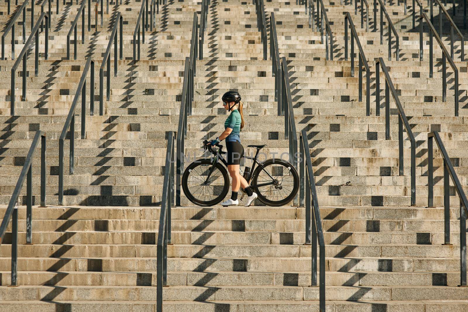 Young professional female cyclist in cycling garment and protective gear standing on the steps with her bike after having a training, riding in city by friendsstock