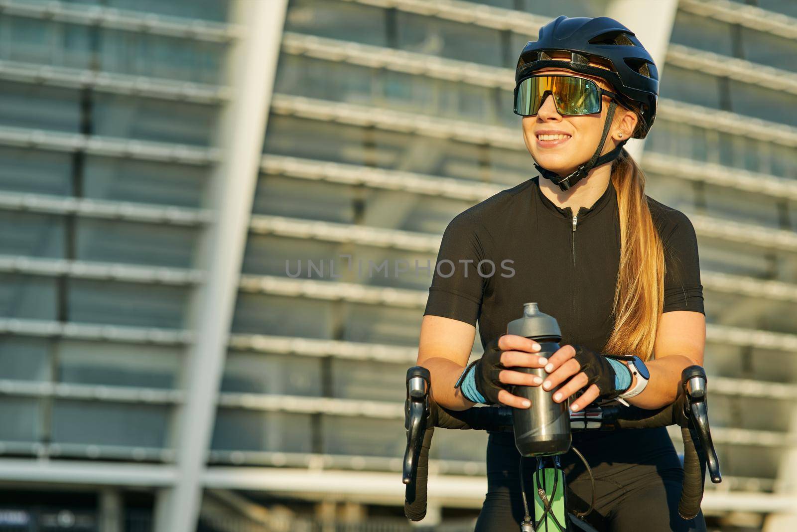 Portrait of smiling sportswoman in black cycling garment and protective gear having a rest while riding bike in city, training outdoors on a sunny day by friendsstock