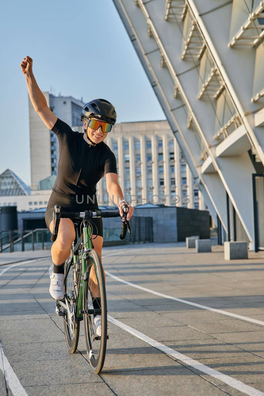 Excited professional female cyclist in black cycling garment and protective gear smiling, raising her arm, looking satisfied with the result of her training by friendsstock
