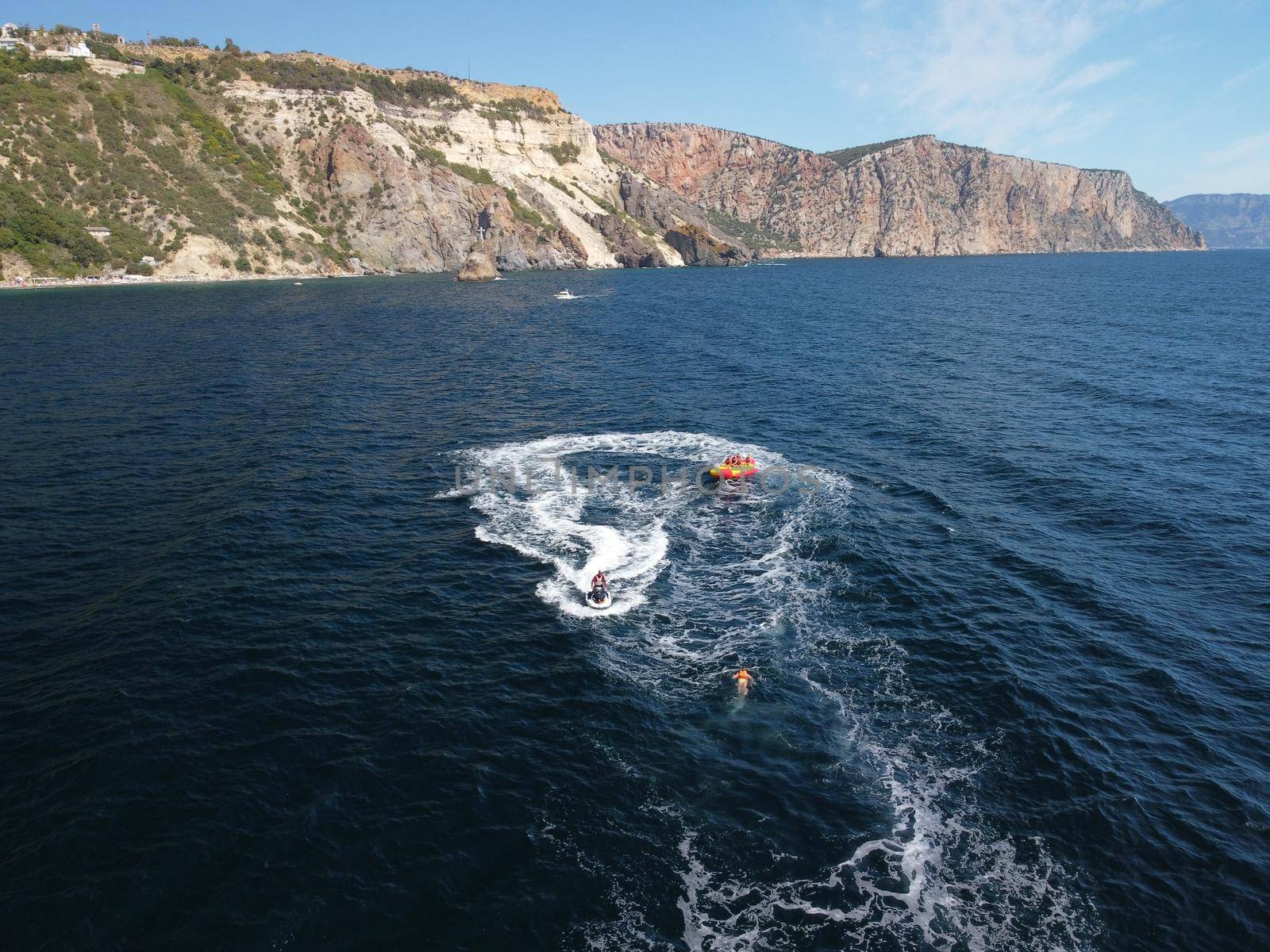 Happy people swim on air mattress behind a high speed water bike. Tourists ride on inflatable watercraft boat. Sea attraction. Water boat skating people on large Inflatable raft. Aerial copter view