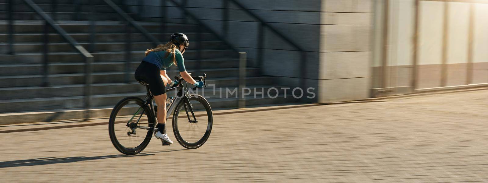 Professional female cyclist in cycling garment and protective gear riding bicycle in city, rushing and passing buildings while training outdoors on a daytime. Urban lifestyle, sports concept