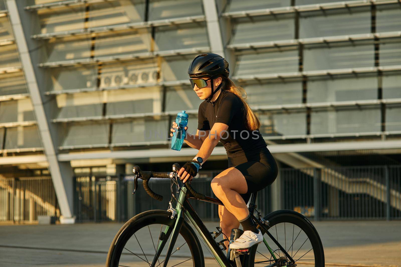 Professional female cyclist in black cycling garment and protective gear holding water bottle while riding bicycle in city, training outdoors at sunset. Urban lifestyle, sports concept