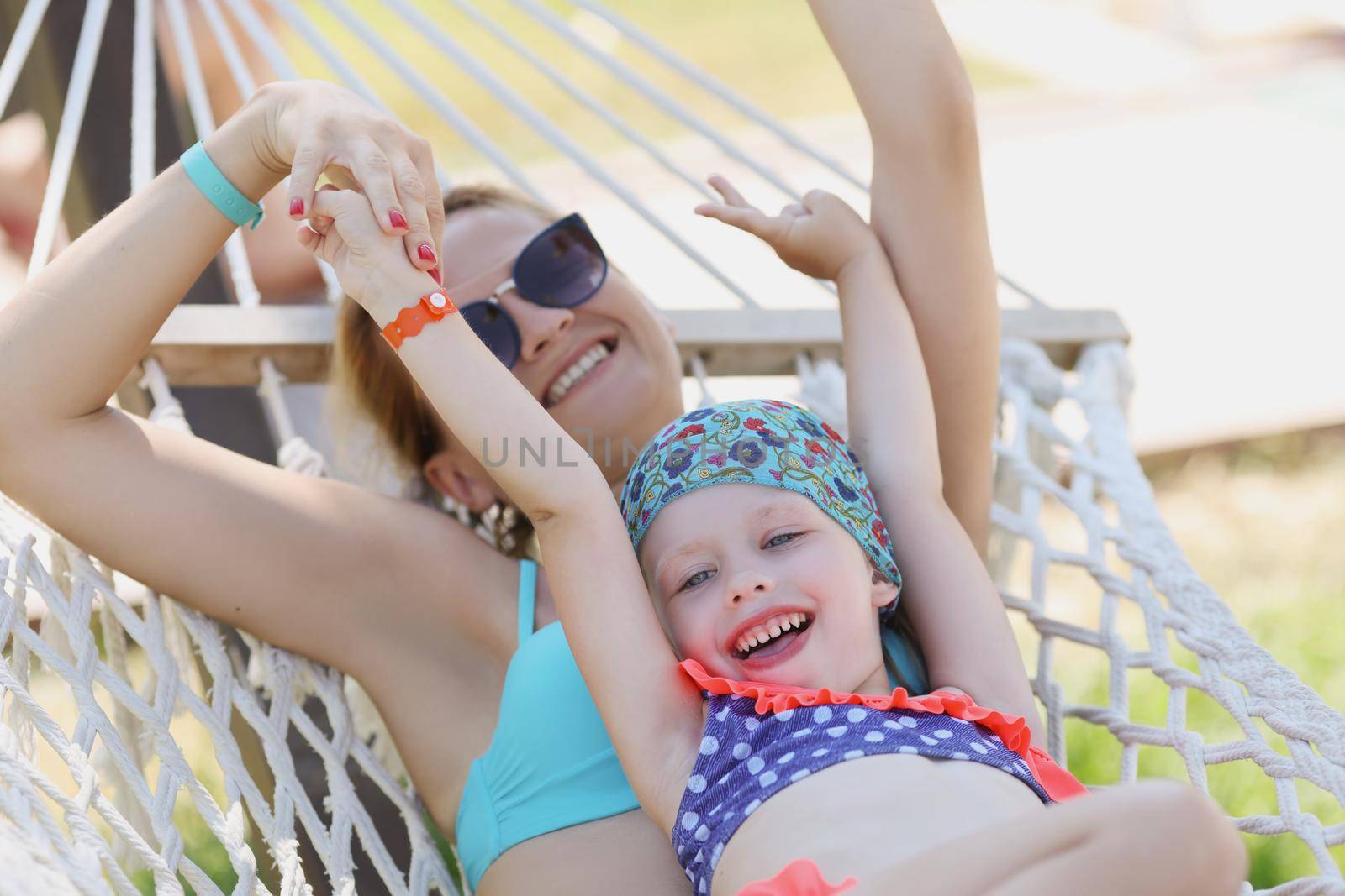 Portrait of happy mother with daughter lay on white hammock in shadow. Girls in swimsuits rest on fresh air on nature. Summertime, quality time concept