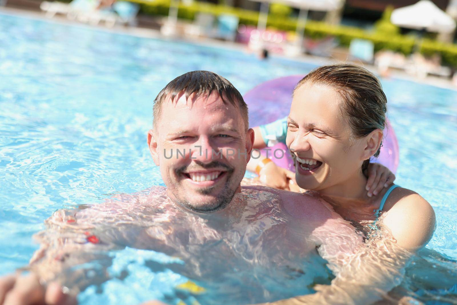 Portrait of cheerful couple posing for picture in swimming pool in luxury resort. Happy laughing woman and smiling husband hugging. Family, summer concept