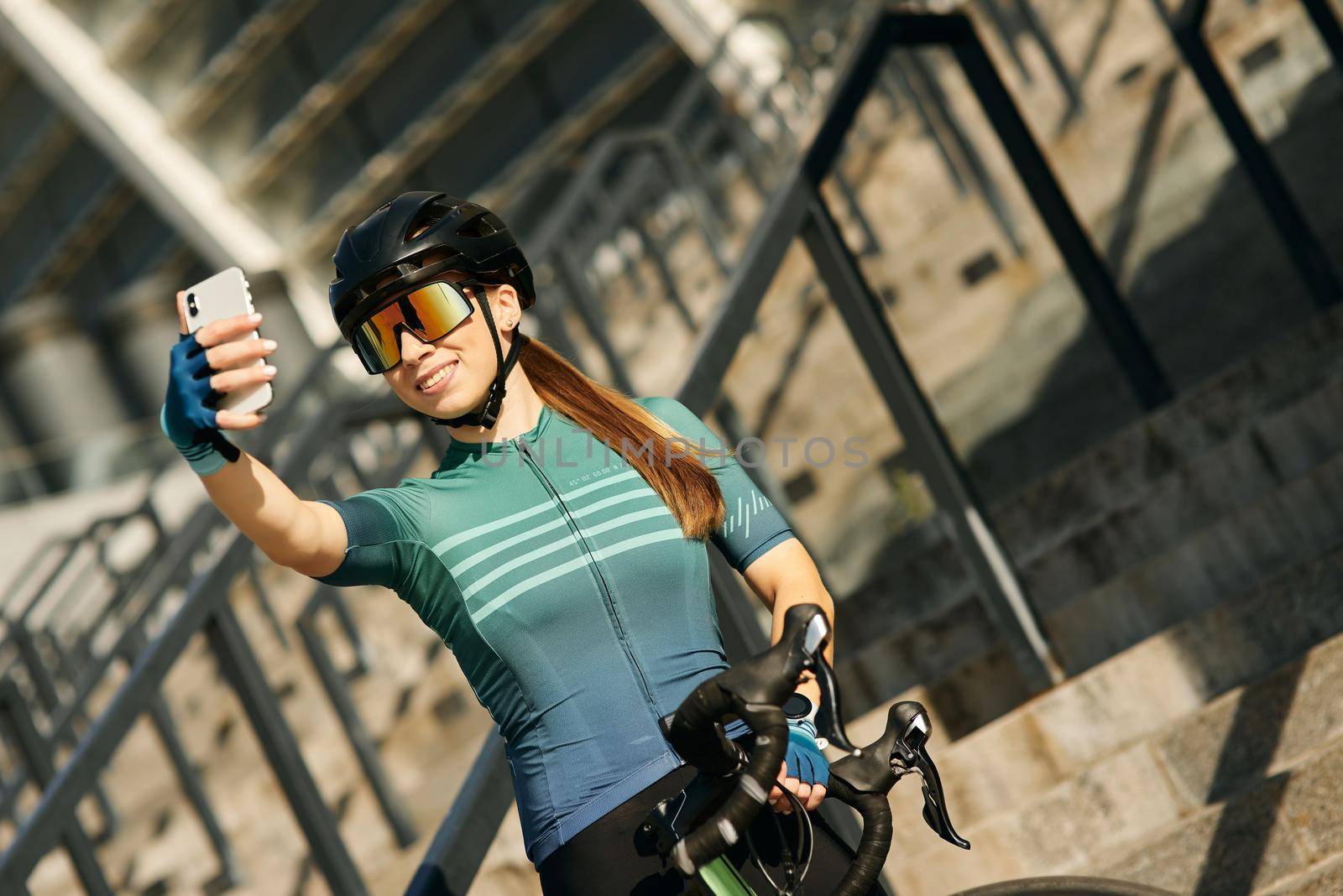 Joyful professional female cyclist in cycling garment and protective gear smiling while taking selfie using smartphone, standing and resting after riding bicycle in city center. Active lifestyle