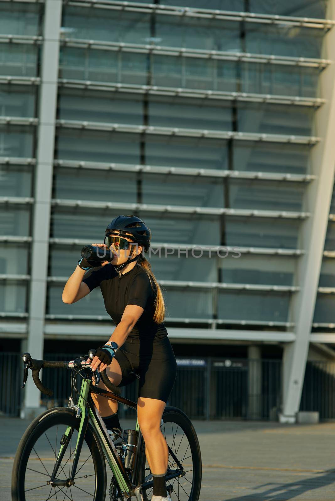 Professional female cyclist in black cycling garment and protective gear drinking water while riding bicycle in city, training outdoors on a warm day. Urban lifestyle, sports concept
