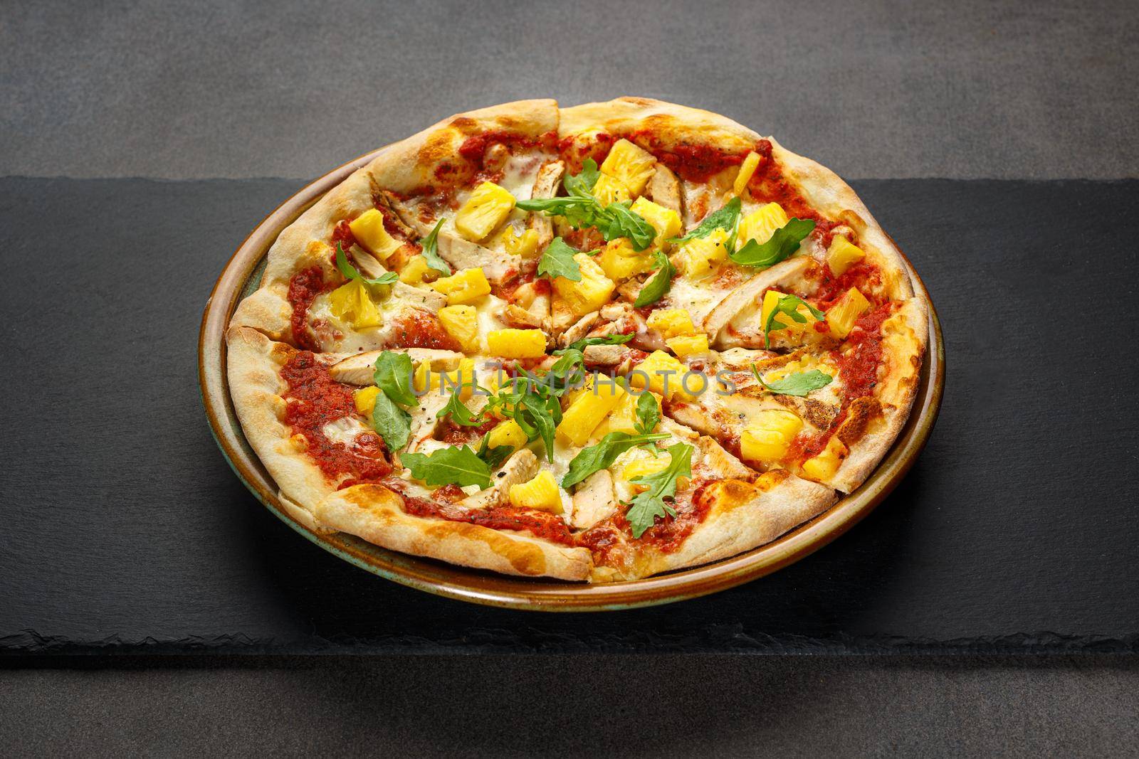 Delicious traditional Italian pizza with pineapple and chicken on on a dark stone board.