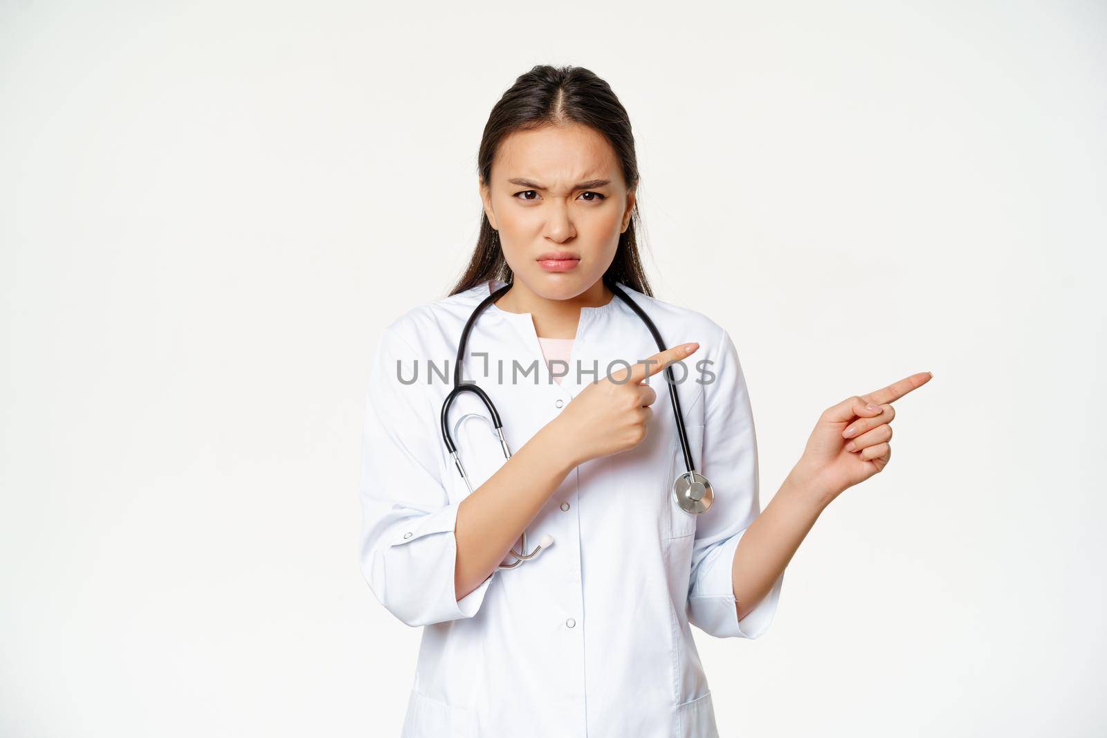Angry female doctor, asian physician in medical robe and stethoscope, pointing fingers right and frowning furious, staring disappointed, white background.