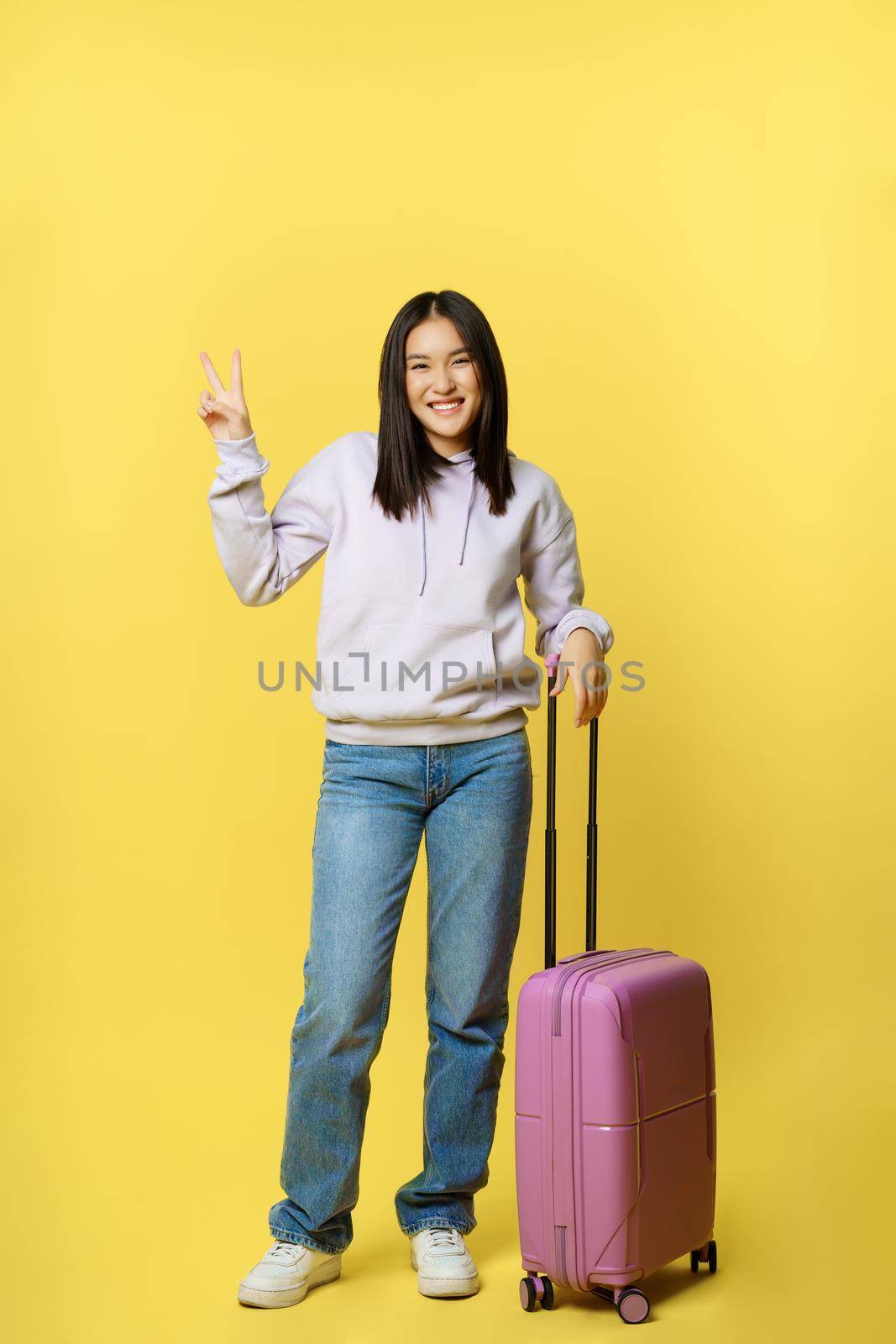 Full length shot happy asian girl, tourist with suitcase going on vacation, smiling and showing peace sign, yellow background.