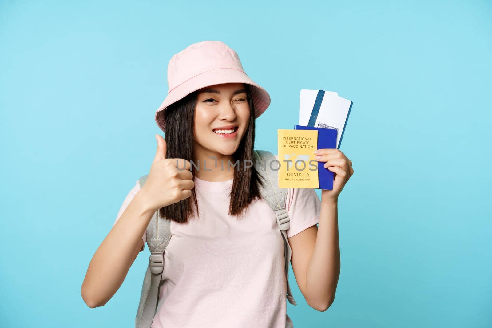 Enthusiastic girl traveller, asian tourist shows thumbs up and international vaccine certificate, health passport and flight tickets, standing over blue background.