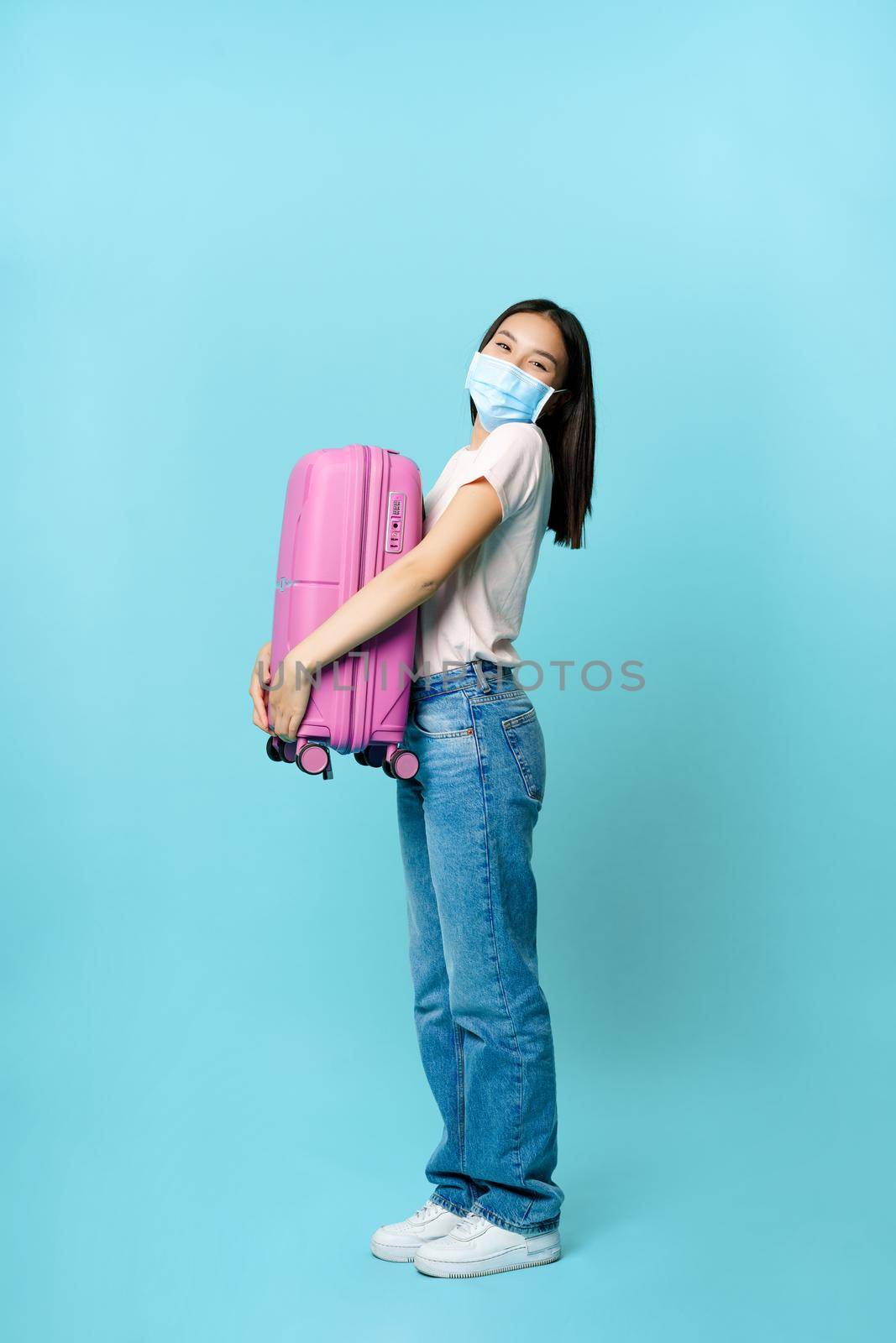 Full length of excited woman tourist, hugging suitcase and smiling, wearing medical face mask from covid-19, travelling abroad, concept of health and tourism.
