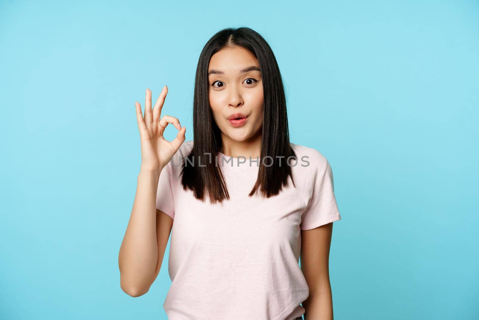Enthusiastic brunette korean girl shows okay sign, watching smth fantastic, awesome price, recommending shop, standing over blue background.