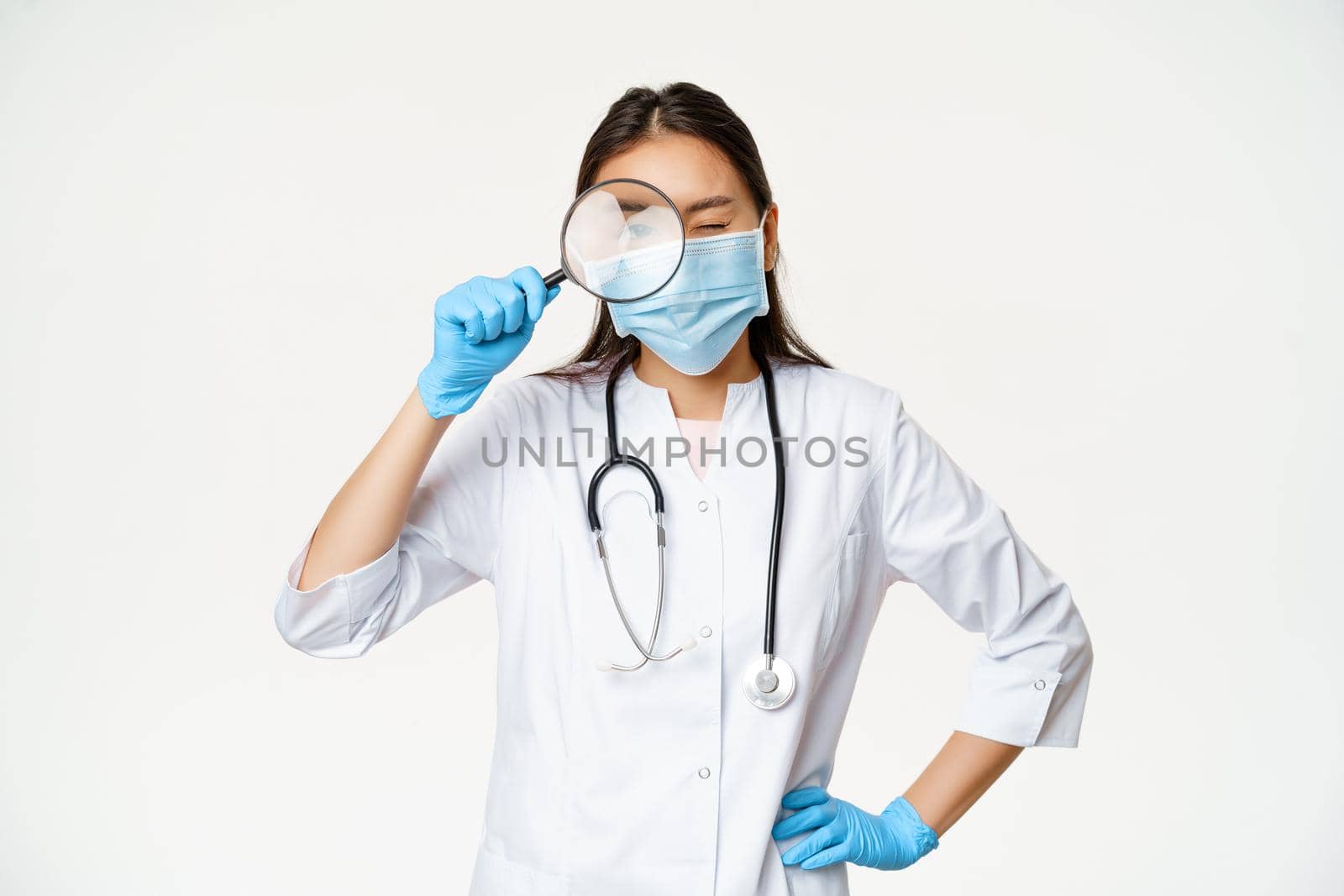Asian female doctor looking through magnifying glass, examine patient, searching disease, wearing medical mask and rubber gloves, white background.
