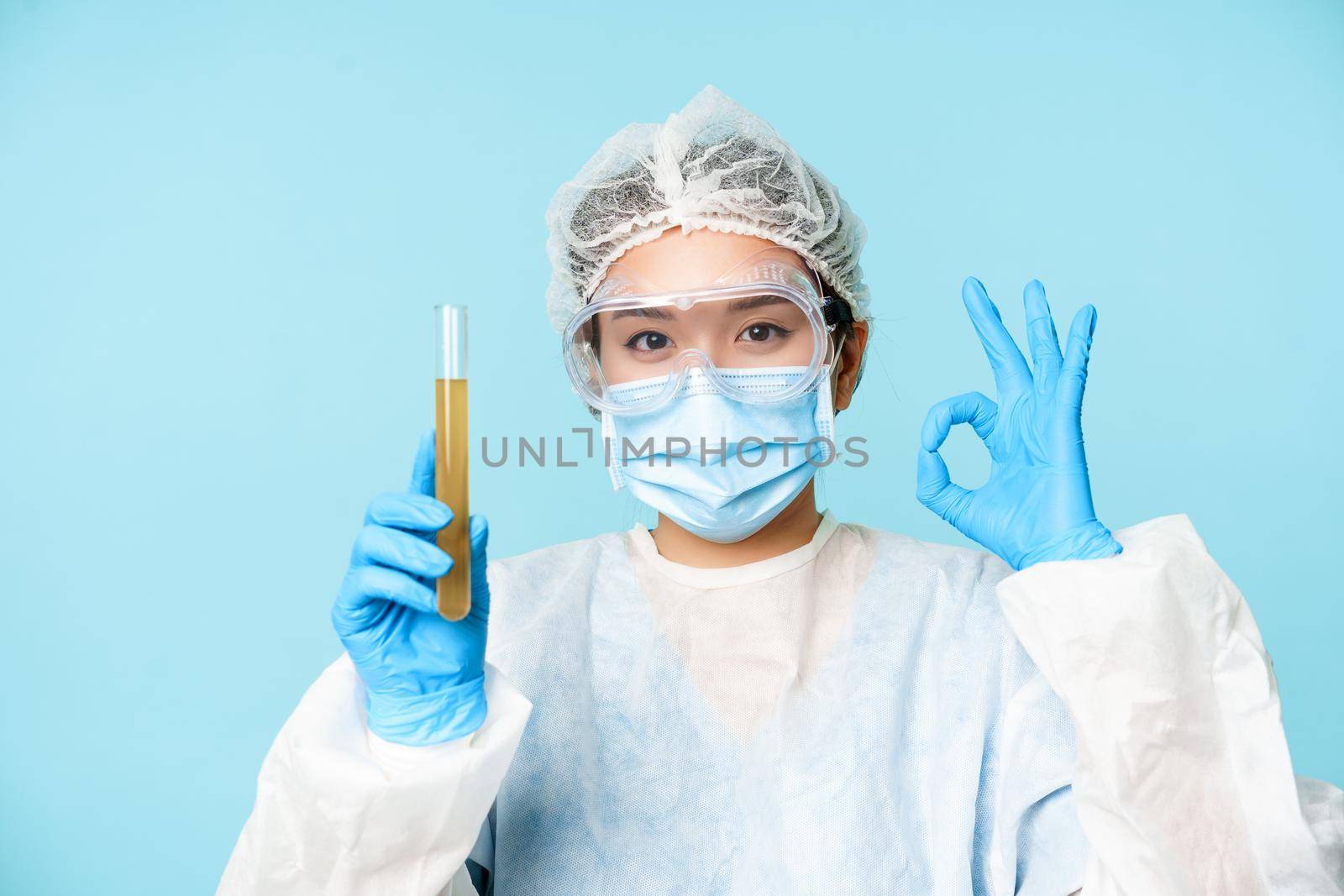 Laboratory worker, nurse in personal protective equipment, showing sample in test tube and okay sign, testing patient analise, blue background.