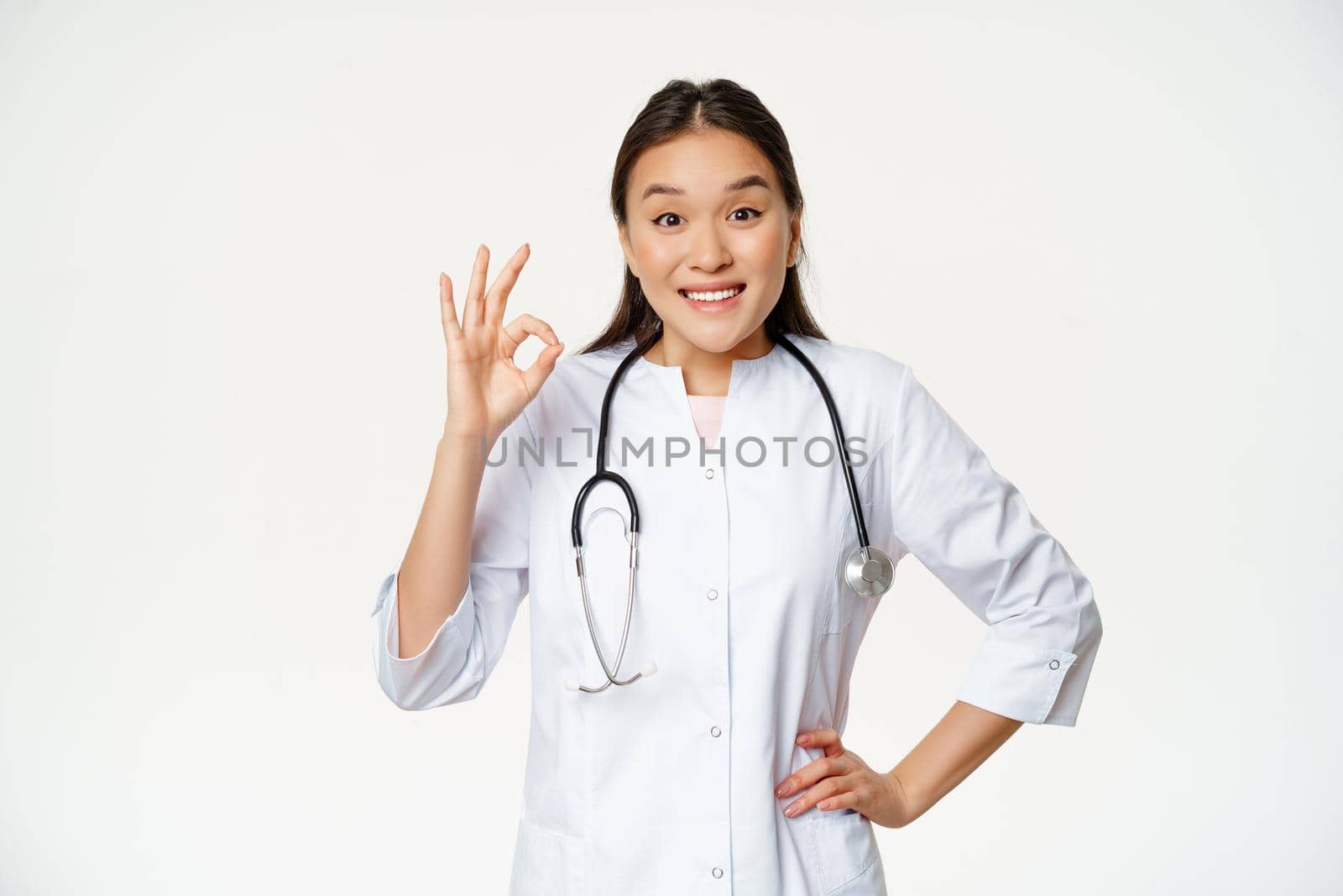 Smiling asian doctor shows okay sign, wears medical robe. Female hospital worker in uniform recommends smth, standing over white background.