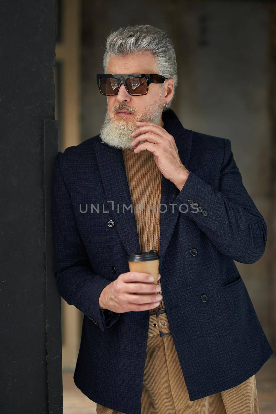 Stylish middle aged man in business casual wear and sunglasses leaning on the wall, holding disposable coffee cup while posing in loft interior by friendsstock