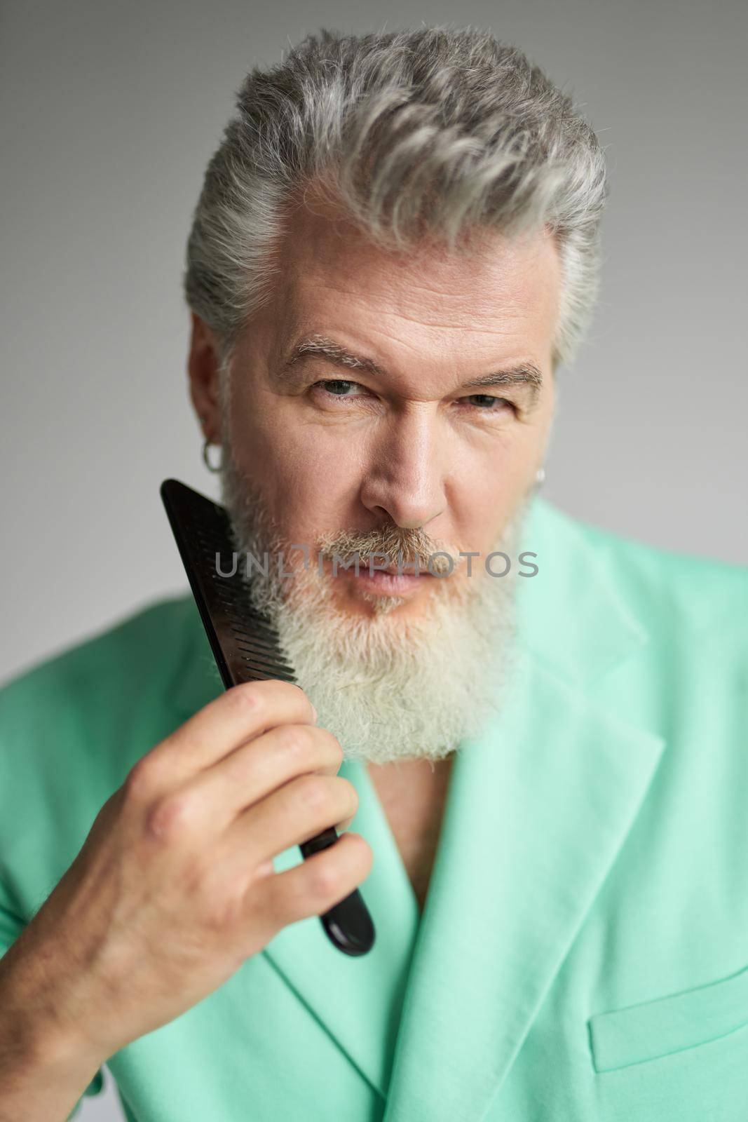 Portrait of attractive gray haired mature man looking at camera, brushing his beard with comb, posing over white background by friendsstock