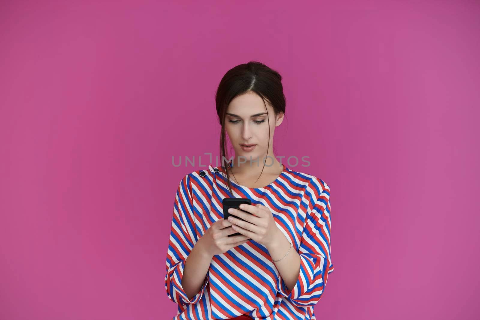 Portrait of young girl with serious face while using smartphone isolated on pink background. Female model chatting or browsing on social networks. Fashion and technology concept