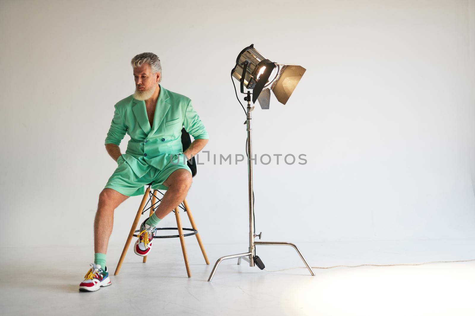 Studio shot of trendy gray haired middle aged man wearing stylish outfit with colorful sneakers looking aside while sitting next to studio spotlight, posing over white background by friendsstock