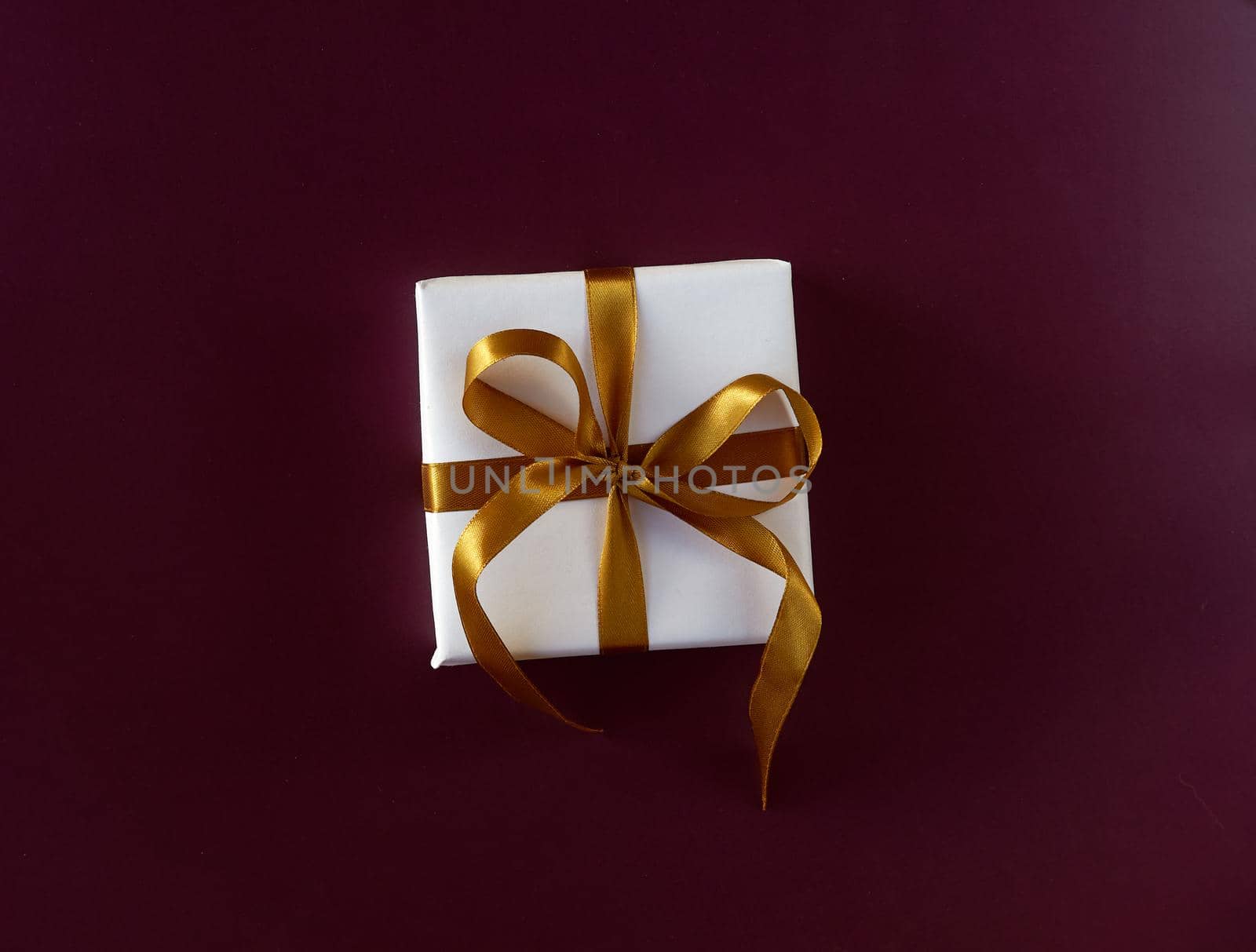 Gift box wrapped in white paper with a golden bow on festive violet background. Copyspace for your text. Flat lay style. Christmas, New Year or birthday celebration concept. Web element for postcard