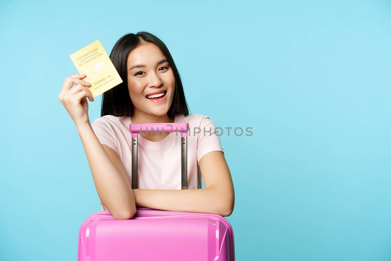 Happy asian girl, tourist with suitcase shows her covid-19 international vaccination passport, getting vaccine to travel abroad during pandemic, blue background.