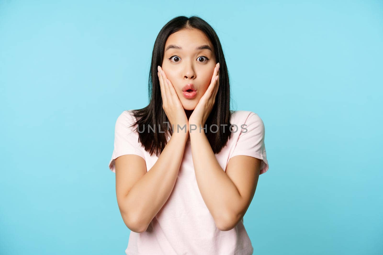 Image of korean woman watching smth amazing with surprised face, looking excited at camera, standing over blue background.
