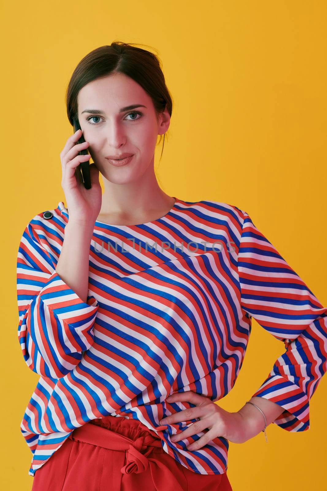 Portrait of young girl talking on the phone while standing in front of yellow background. Female model representing modern fashion and technology concept