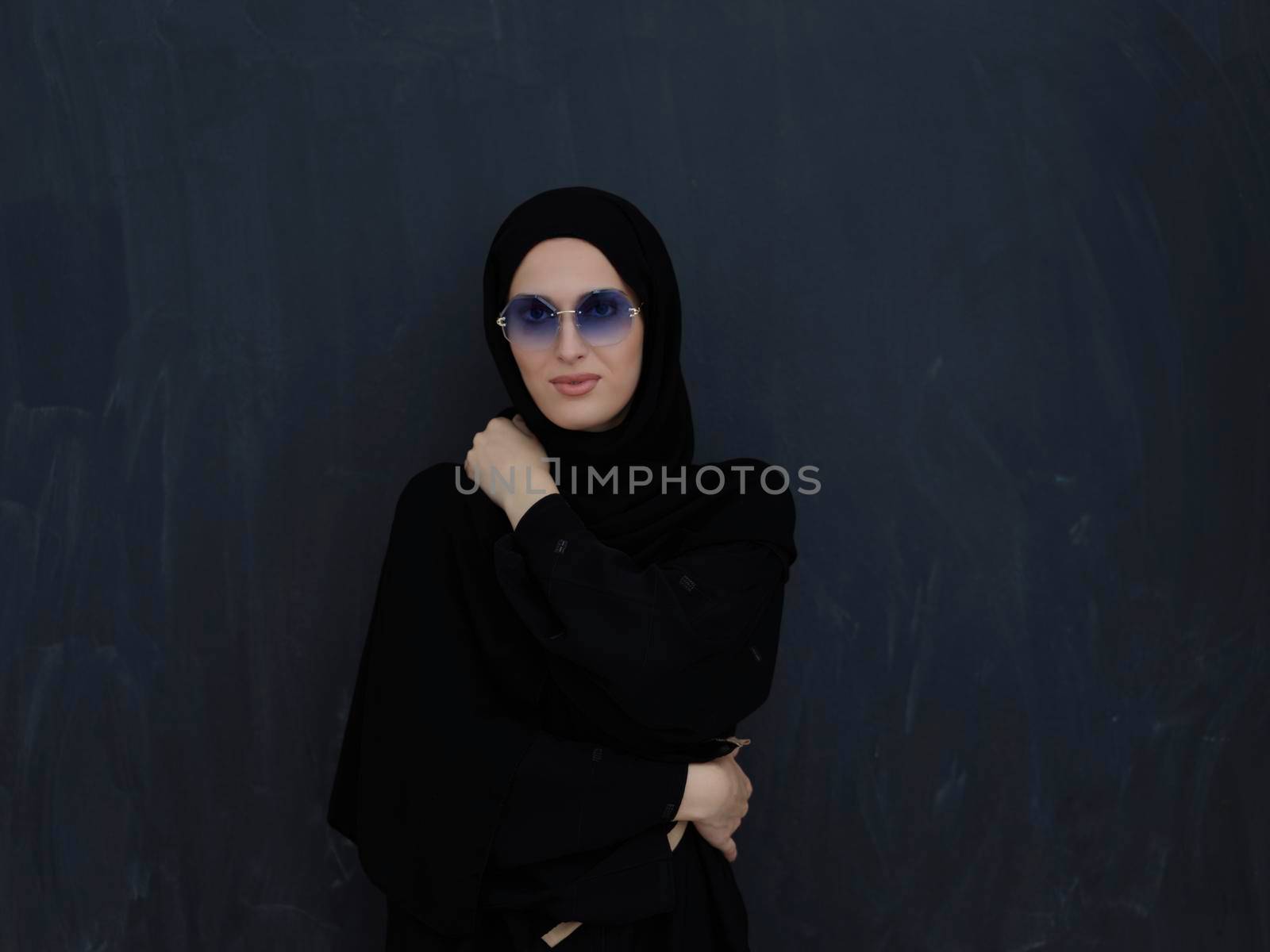 Young muslim in traditional clothes or abaya and sunglasses posing in front of black chalkboard. Arab woman representing modern arabic lifestyle, islamic  fashion and Ramadan kareem concept