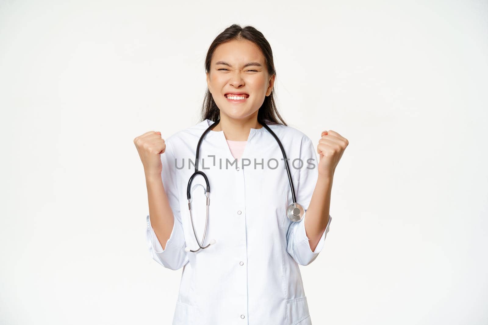 Happy asian intern celebrating. Physician doctor triumphing and rejoicing, say yes with clenched fists pump, standing ecstatic over white background.