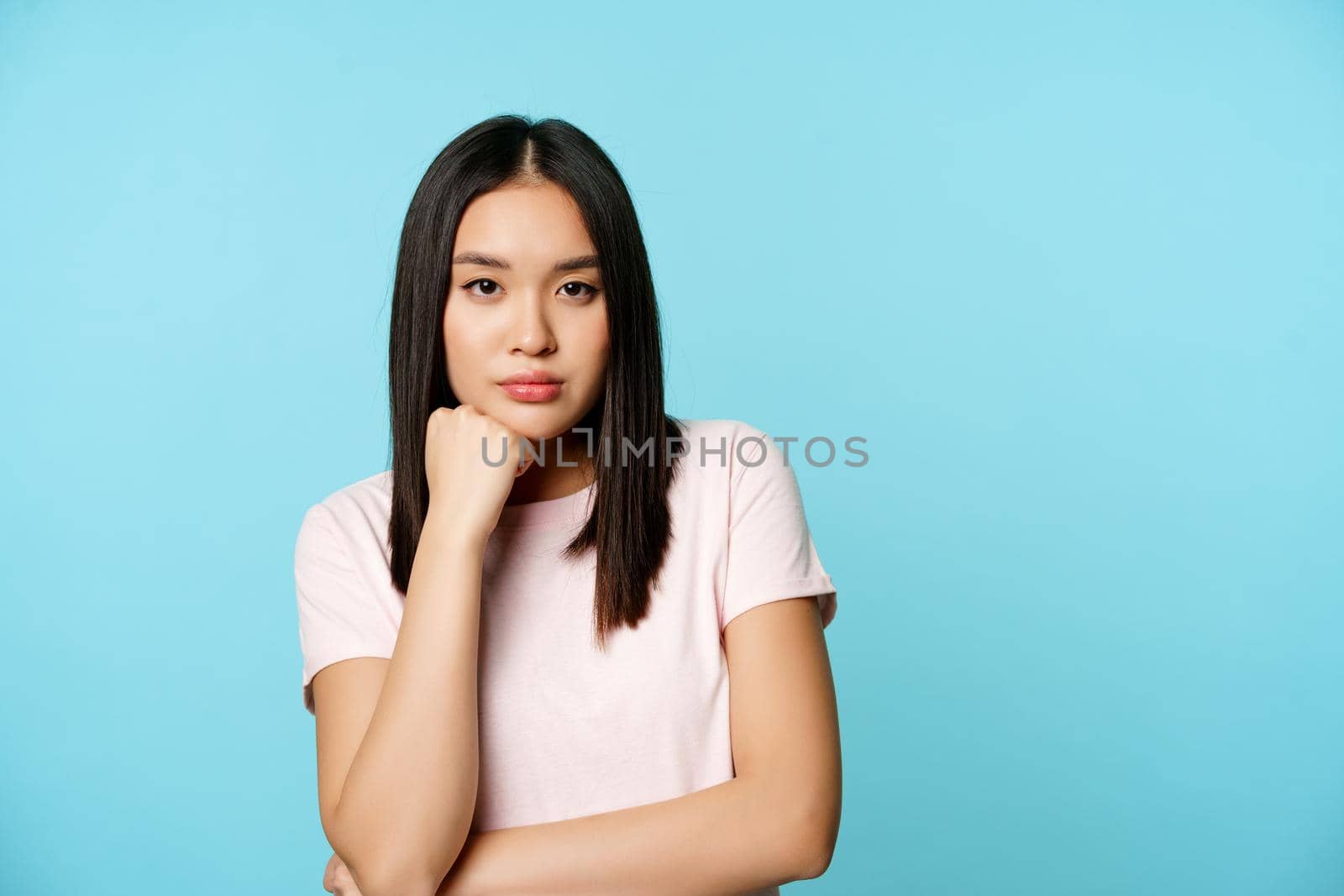 Asian girl watching smth boring, looking with unamused, reluctant face expression, standing in t-shirt over blue backround by Benzoix