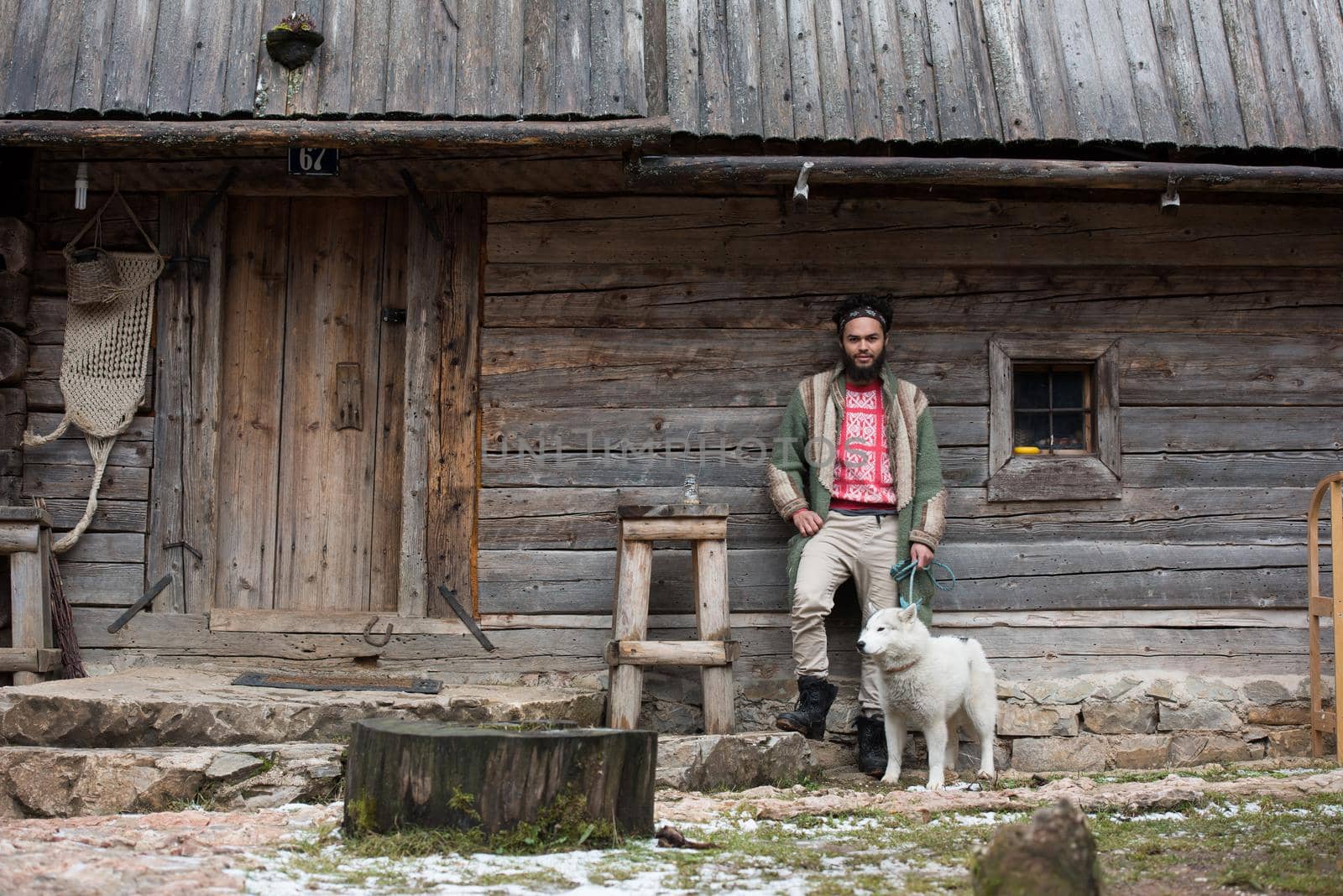 handsome young hipster man standing together with white husky dog in front of old vintage retro wooden house