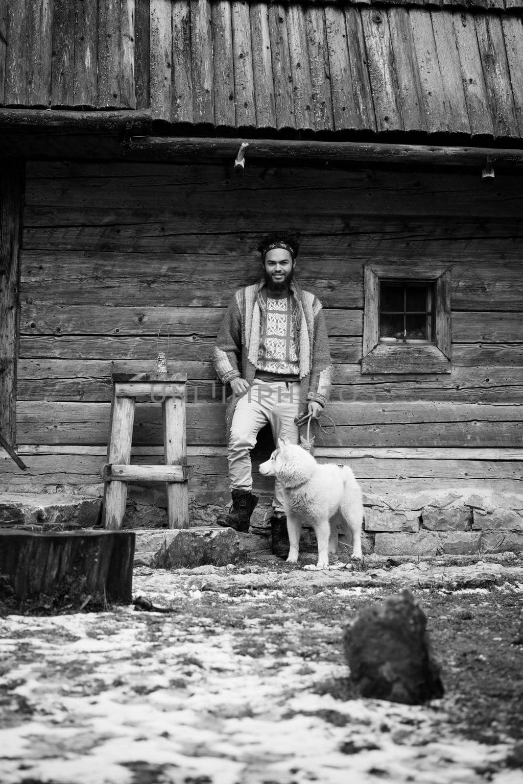 handsome young hipster man standing together with white husky dog in front of old vintage retro wooden house