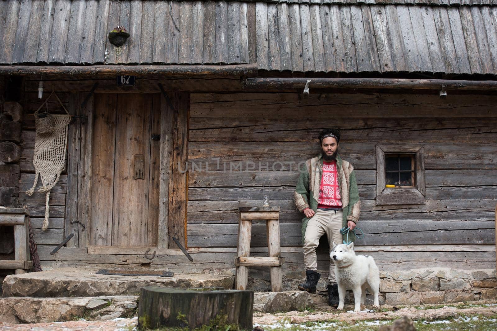 handsome young hipster man standing together with white husky dog in front of old vintage retro wooden house