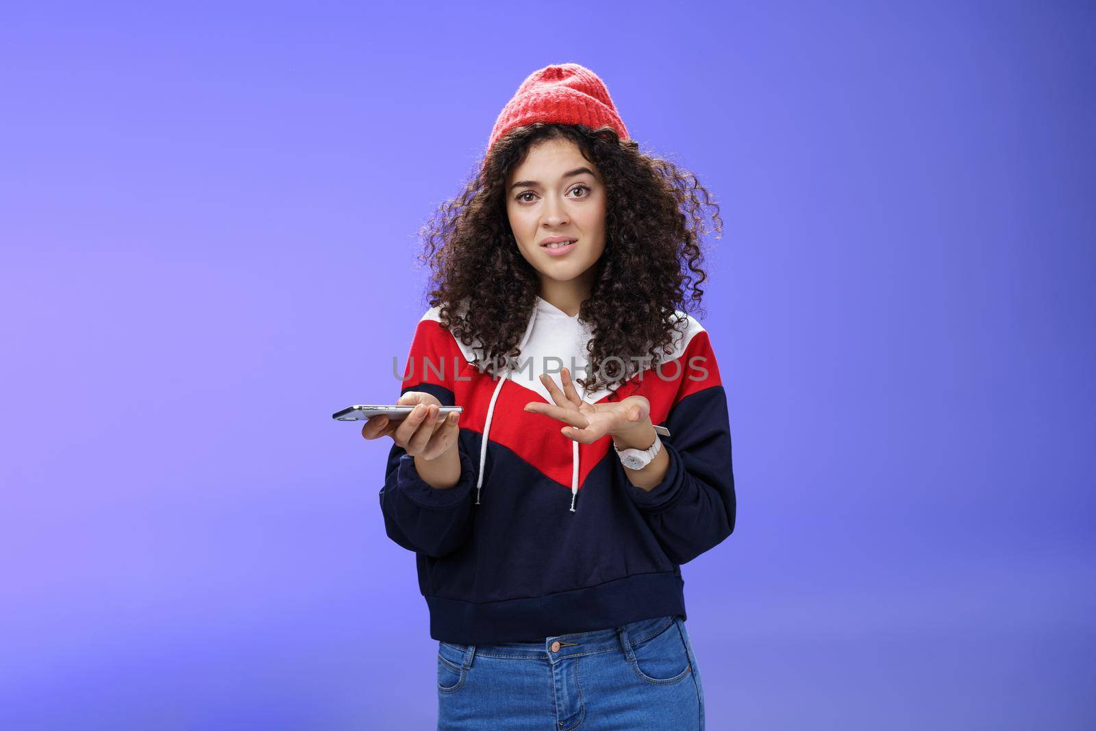 Confused and clueless girl cannot understand meaning of strange message shrugging grimacing questioned as holding smartphone and extending arm in unaware gesture over blue background by Benzoix