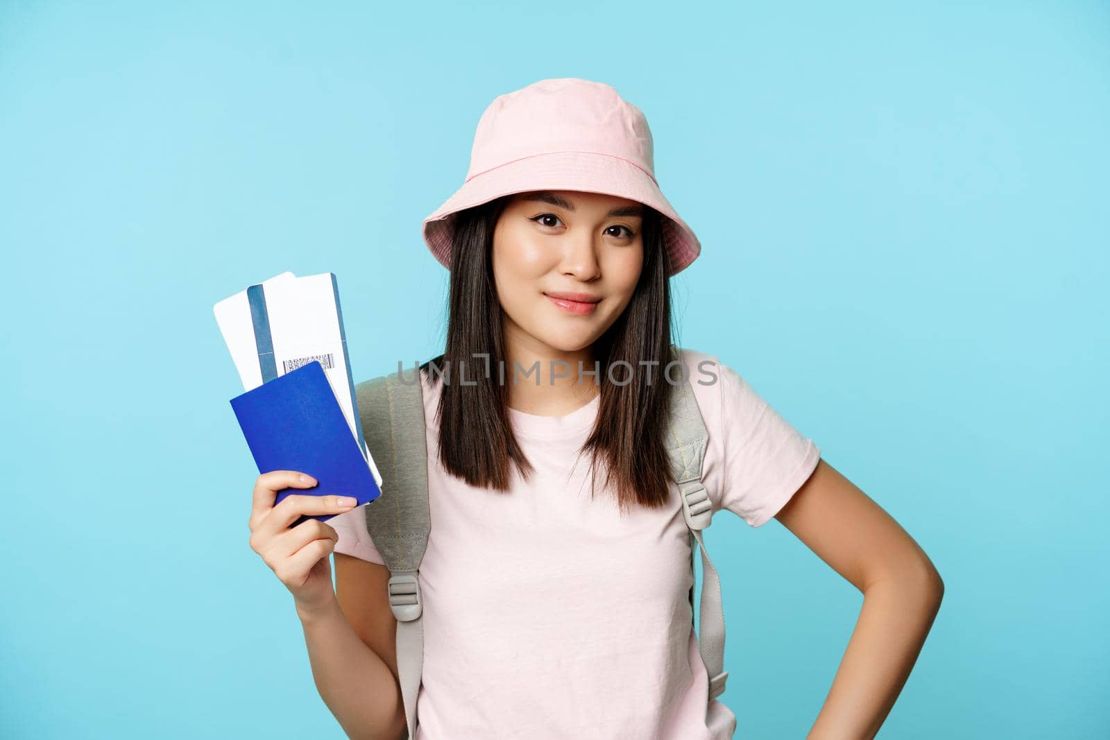 Smiling korean girl tourist, showing passport and tickets for flight, booking hotel, going on trip, voyage, standing over blue background.