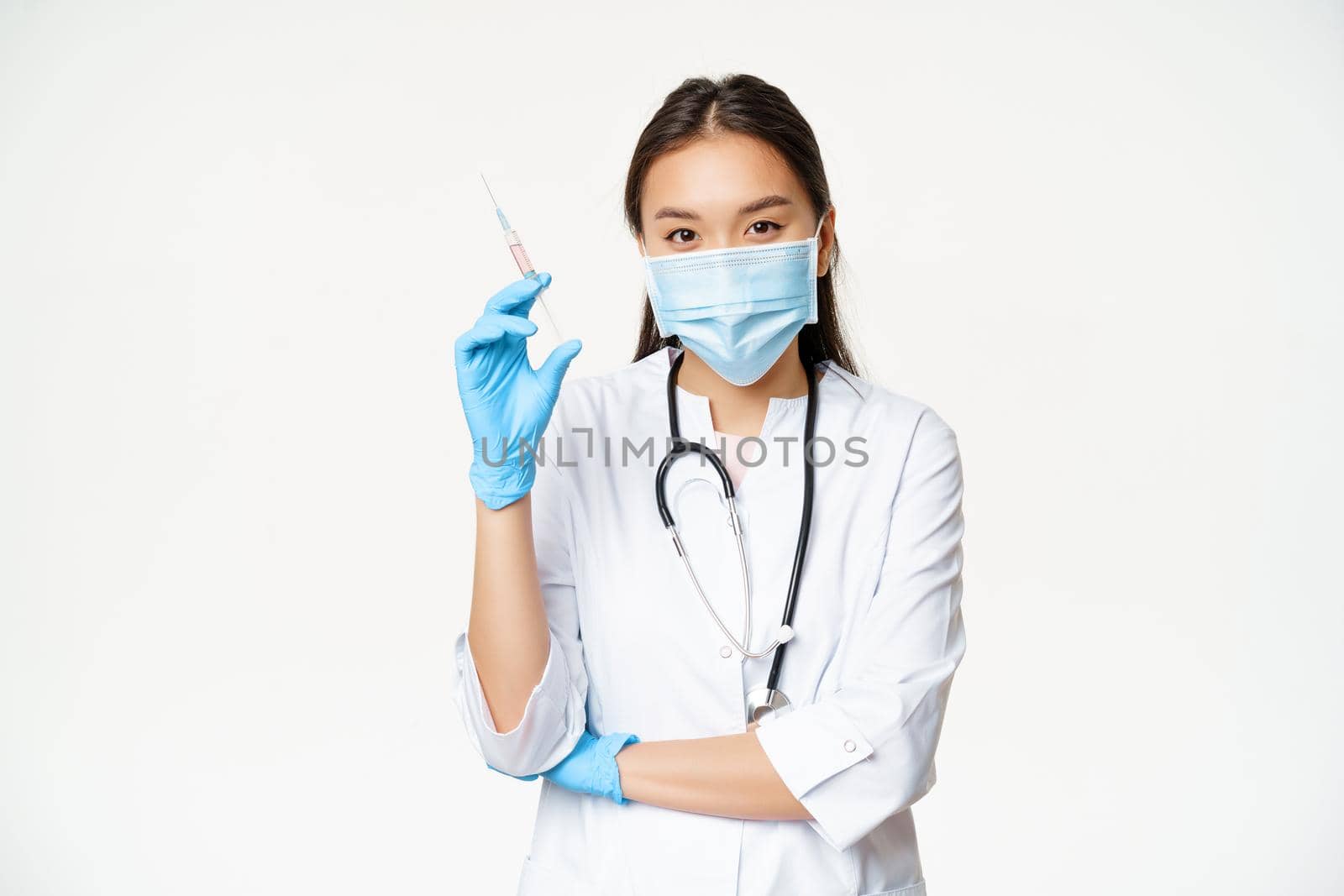 Vaccination and healthcare concept. Asian female doctor, nurse in medical face mask and gloves holding syringe with vaccine, white background.