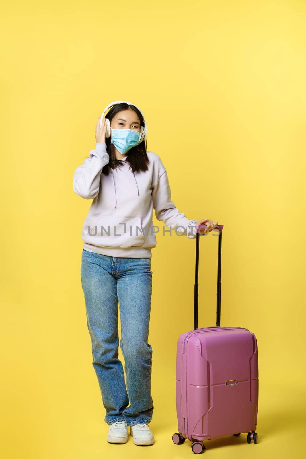 Full length shot happy asian young girl tourist, international traveller in headphone, standing near suitcase and smiling, wearing medical face mask from covid-19 disease by Benzoix