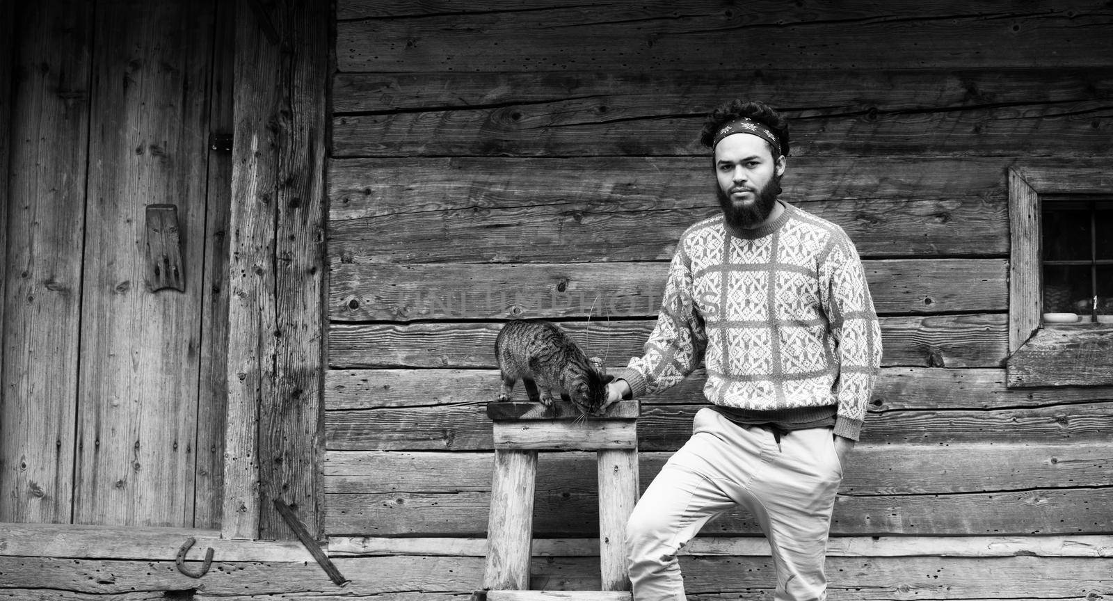 portrait of young hipster man with beard and cat in front of wooden house